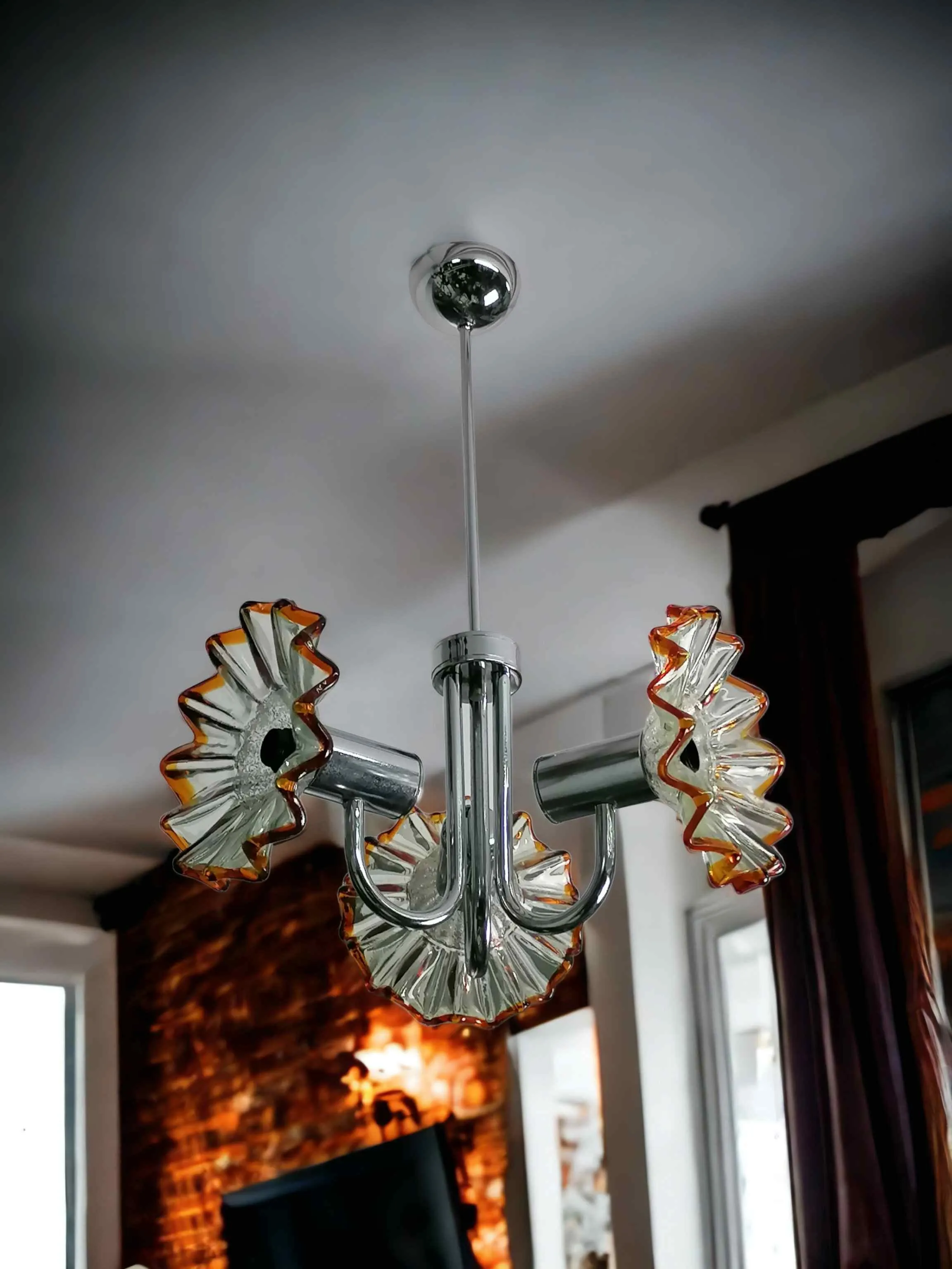 Ce lustre Murano de Mazzega incarne l'esprit élégant et ludique du design italien des années 70. Réalisé en verre soufflé de Murano, il présente trois bras en métal chromé supportant autant d'abat-jours en verre plissé avec des détails en nuances orangées, typiques du mouvement Space Age de l'époque. Les formes audacieuses et les finitions chromées confèrent à cette lampe un charme rétro et sophistiqué, parfait pour ajouter une touche de vintage chic à n'importe quel environnement. Idéal pour les amateurs de design d'auteur, cette lampe est plus qu'un simple accessoire : c'est un véritable morceau d'histoire du design italien.