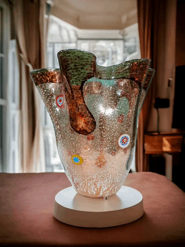 Le vase de Murano Mouchoir Carlo Nason est une œuvre distinctive qui célèbre l'art du verre de Murano, réalisée par le célèbre designer Carlo Nason. Ce vase, signé par le maître, est un exemple exceptionnel de design innovant et de tradition artisanale. Son nom, Mouchoir, vient de la forme qui évoque un mouchoir de tissu flottant, plié et moulé dans le verre, avec des lignes douces et fluides créant un effet dynamique. Caractéristiques principales : Design Unique : Le vase Mouchoir se distingue par sa forme élégante et sinueuse, qui semble presque danser dans l'air. Le pli du verre évoque le mouvement et la légèreté, rendant chaque exemplaire unique. Technique Muranaise : Réalisé à la main selon la tradition verrière de Murano, ce vase est le fruit d'une fabrication artisanale impliquant des techniques de soufflage et de modelage du verre, garantissant un produit final de haute qualité et riche en détails. Couleur et Transparence : Les vases de Carlo Nason sont souvent caractérisés par des couleurs vibrantes et une grande transparence, jouant de manière spectaculaire avec la lumière. Le Mouchoir ne fait pas exception, créant un contraste fascinant entre ses plis doux et les couleurs choisies. Pourquoi le choisir : Le vase de Murano Mouchoir Carlo Nason est parfait pour ceux qui souhaitent un élément décoratif alliant modernité et tradition. C'est une œuvre qui enrichit non seulement l'espace avec son esthétique unique, mais représente également un investissement dans une œuvre d'art verrière de haute classe. Sa forme élégante et son design contemporain en font l'objet idéal pour les intérieurs modernes ou comme complément de luxe dans des environnements plus classiques.